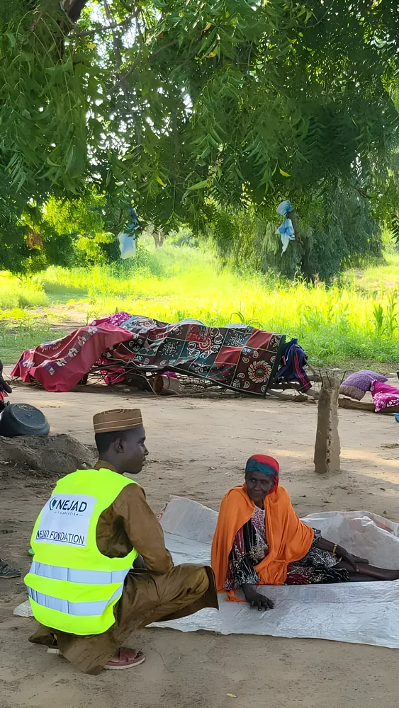 An elderly woman sits on a mat in the open, her belongings piled behind her, as a NEJAD volunteer listens