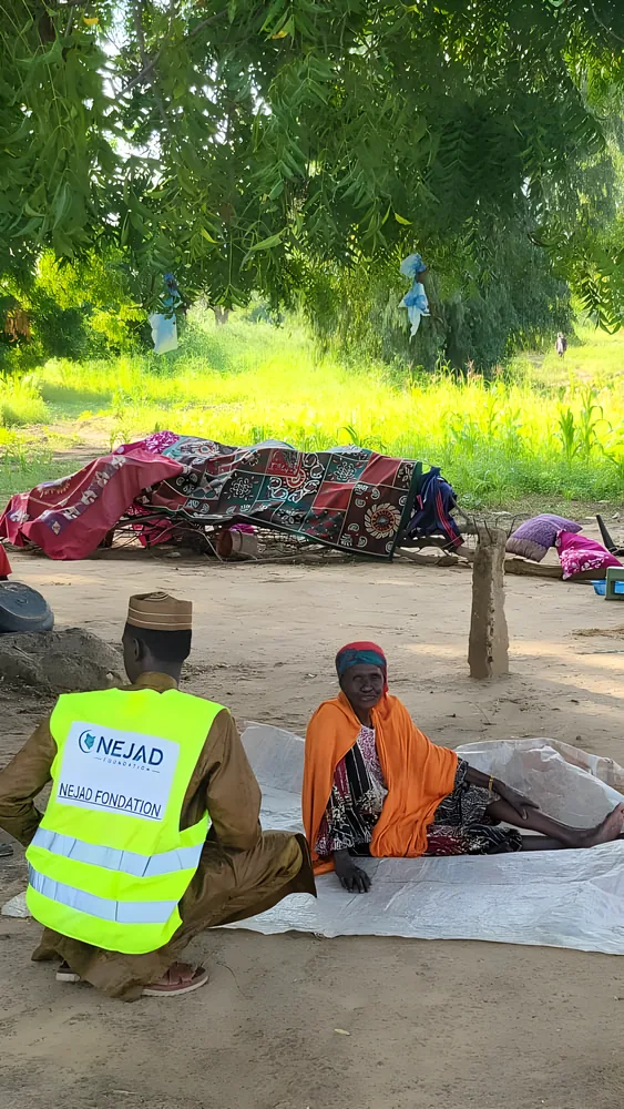 A NEJAD volunteer sits with an elderly woman beside her belongings under a tree