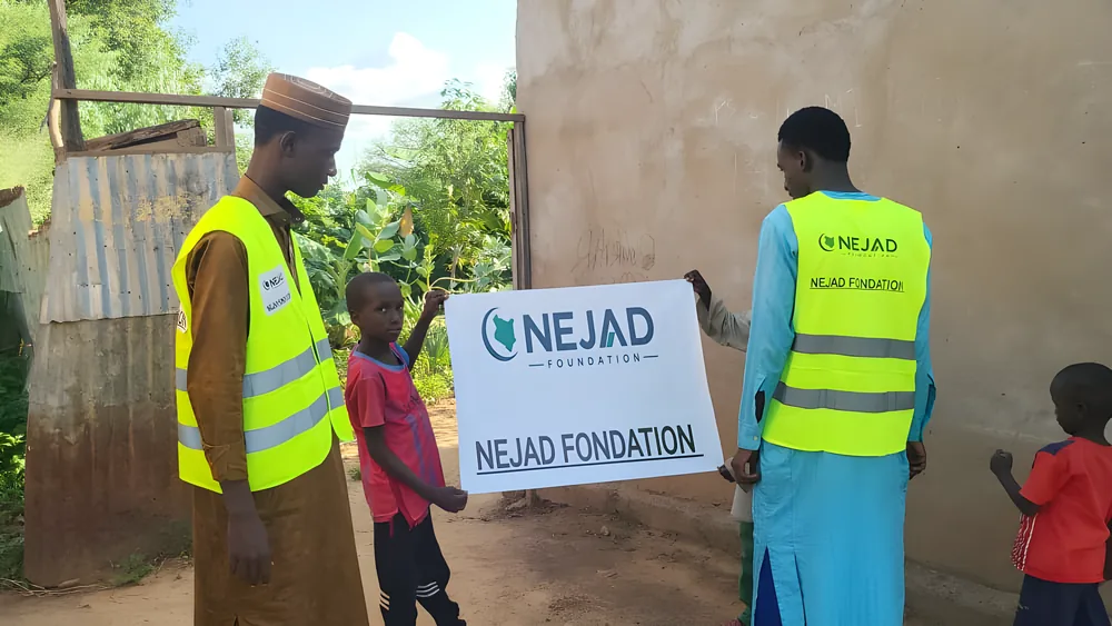 NEJAD volunteers with children beside a village wall, holding the NEJAD banner